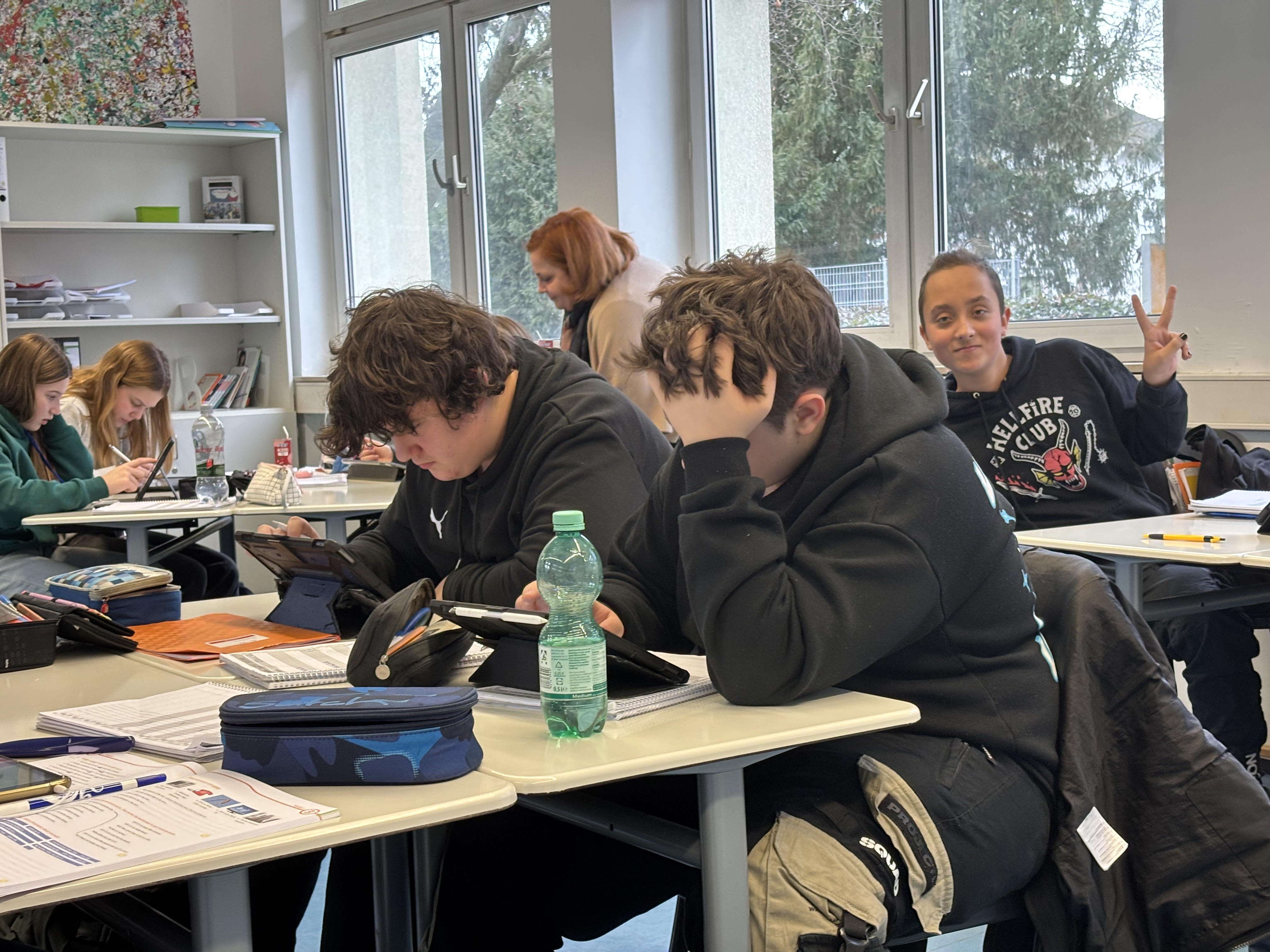 Schüler in einem Klassenzimmer, über ihre Hefte gebeugt, im Hintergrund blickt ein Junge auf und macht das Peace-Zeichen.