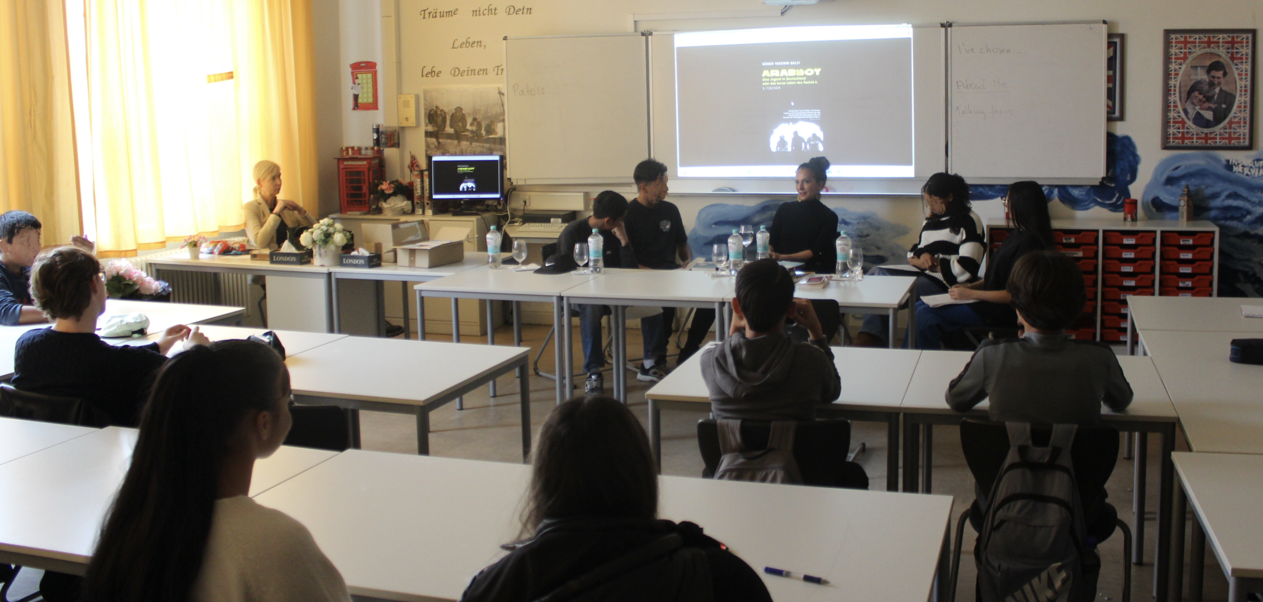 Klassenzimmer mit Schuelerinnen und Schuelern, vorn an der Tafel "Arabboy"-Autorin Güner Yasemin Balci.