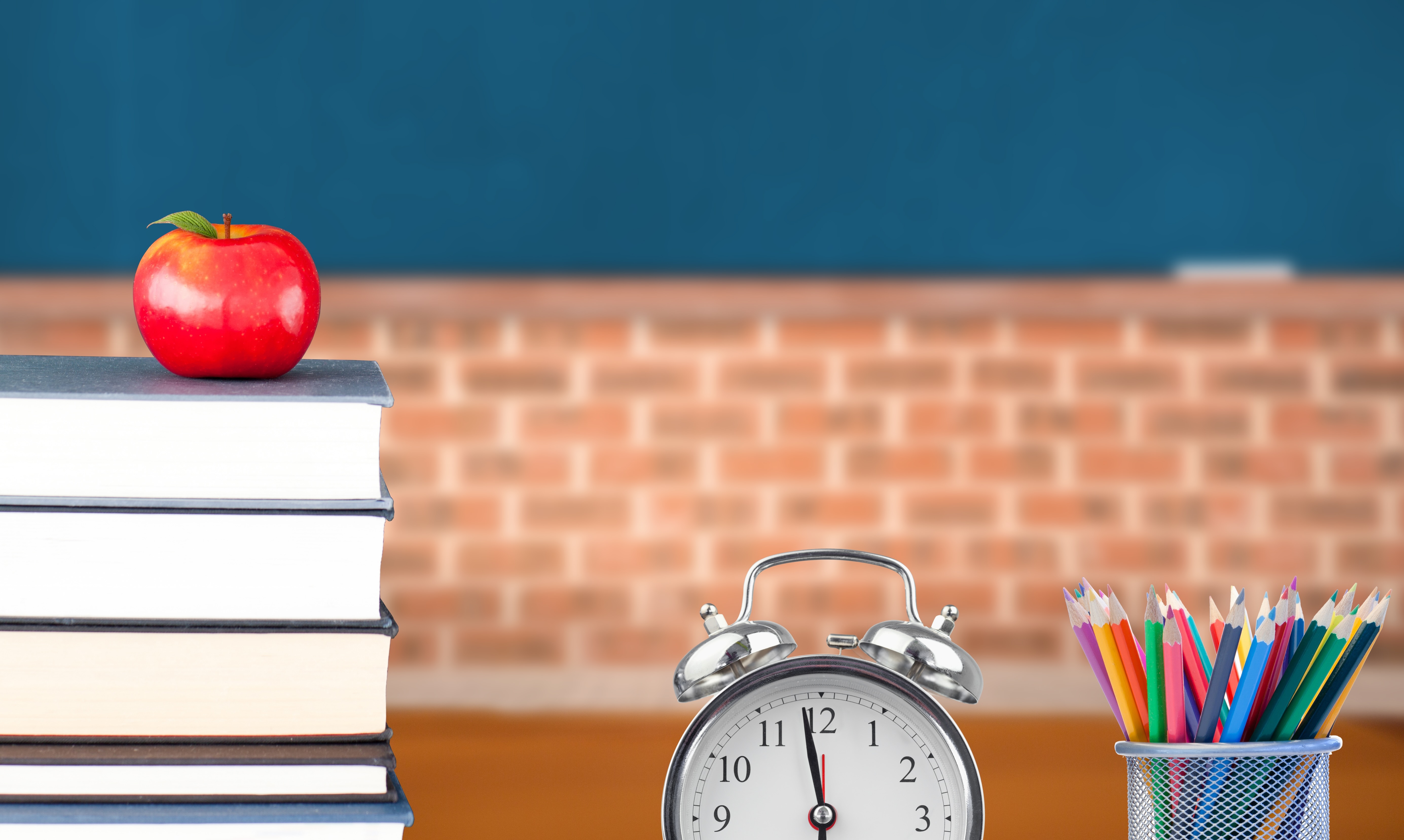Stilleben Lehrerschreibtisch mit Buechern, Apfel, Wecker und Becher mit Buntstiften.