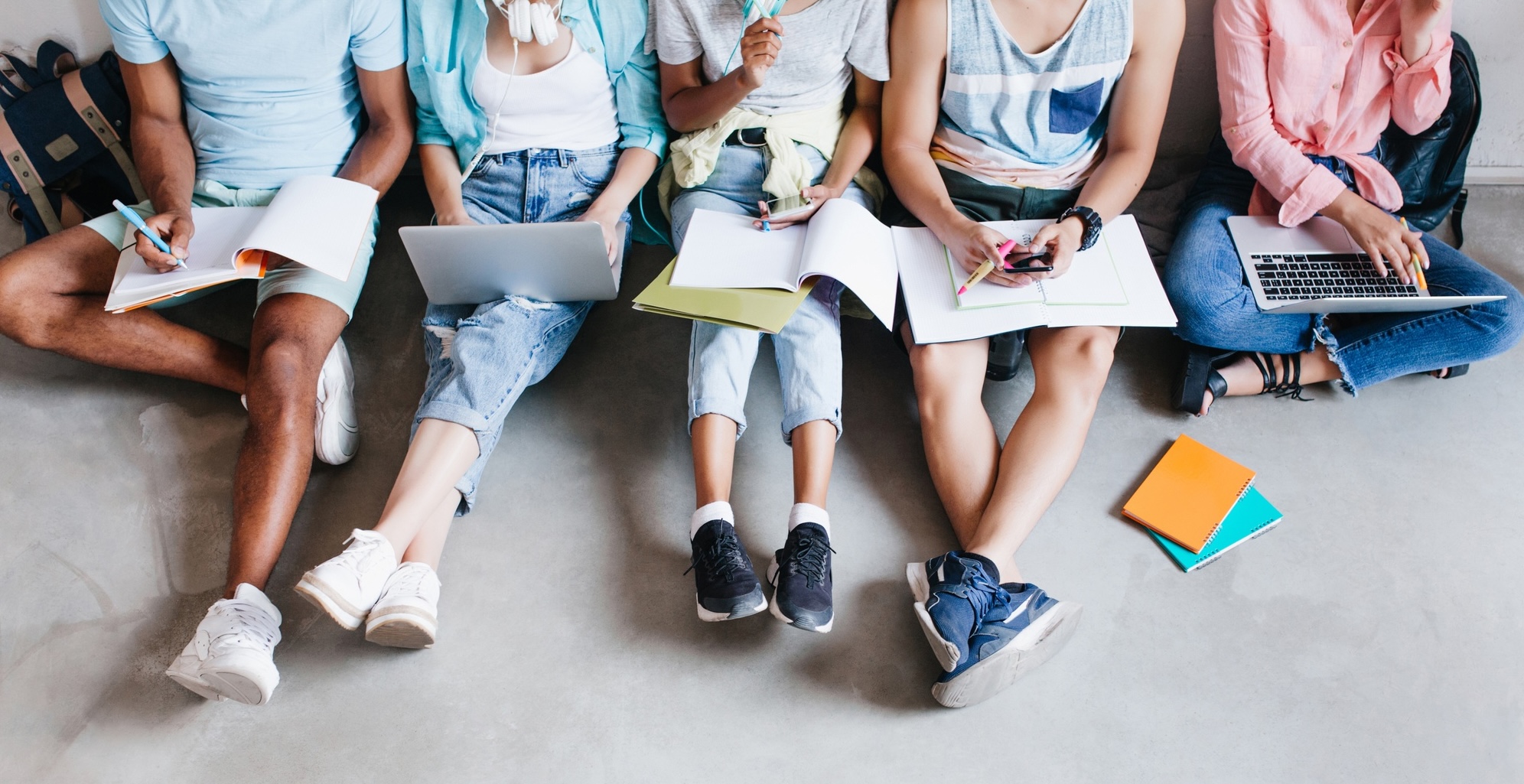 Gruppe junger Menschen auf dem Boden sitzend mit Buechern, Heften, Laptops und Smartphones.