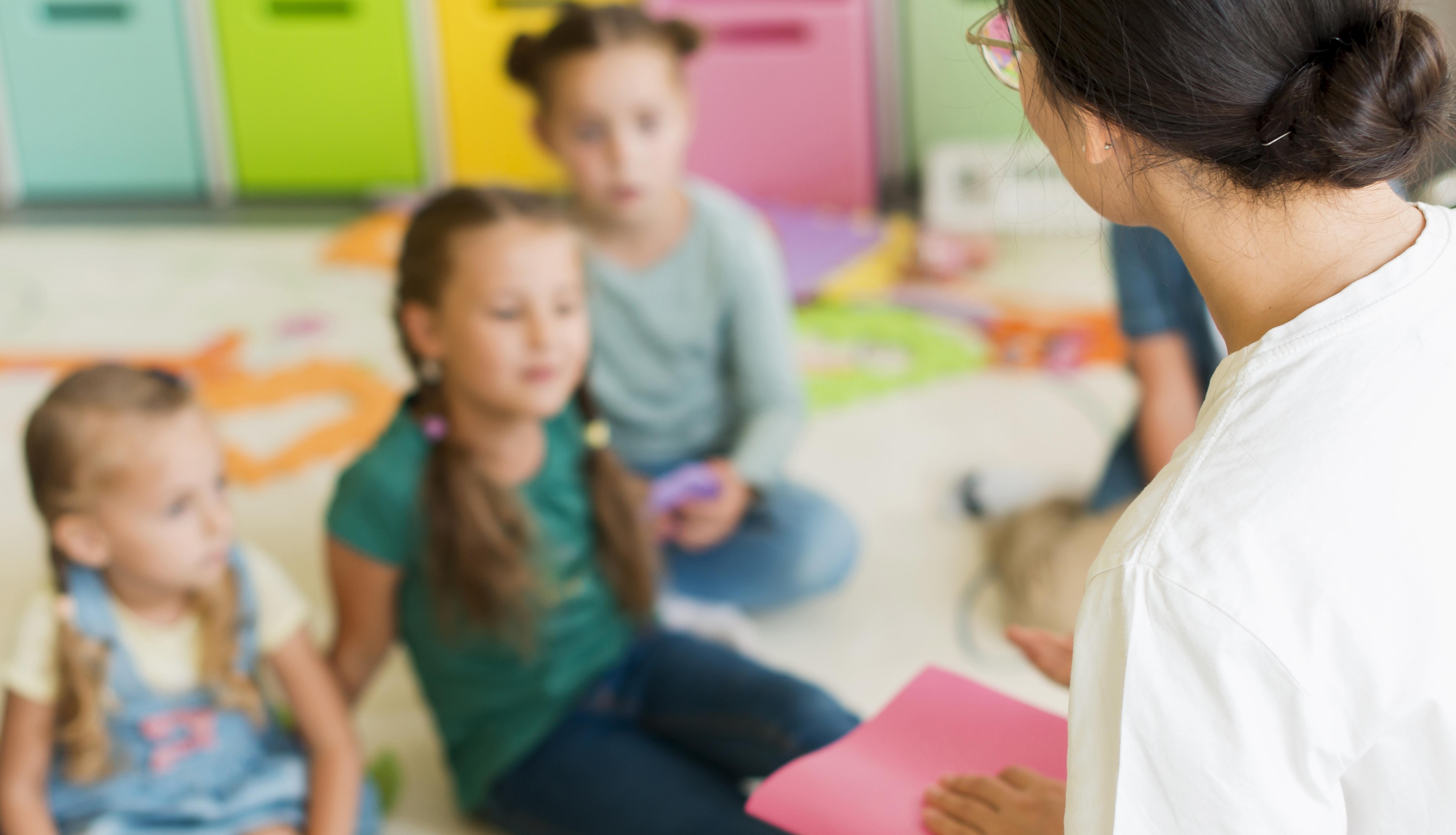 Drei Kindergartenkinder sitzen vor der Erzieherin und hören ihr zu.