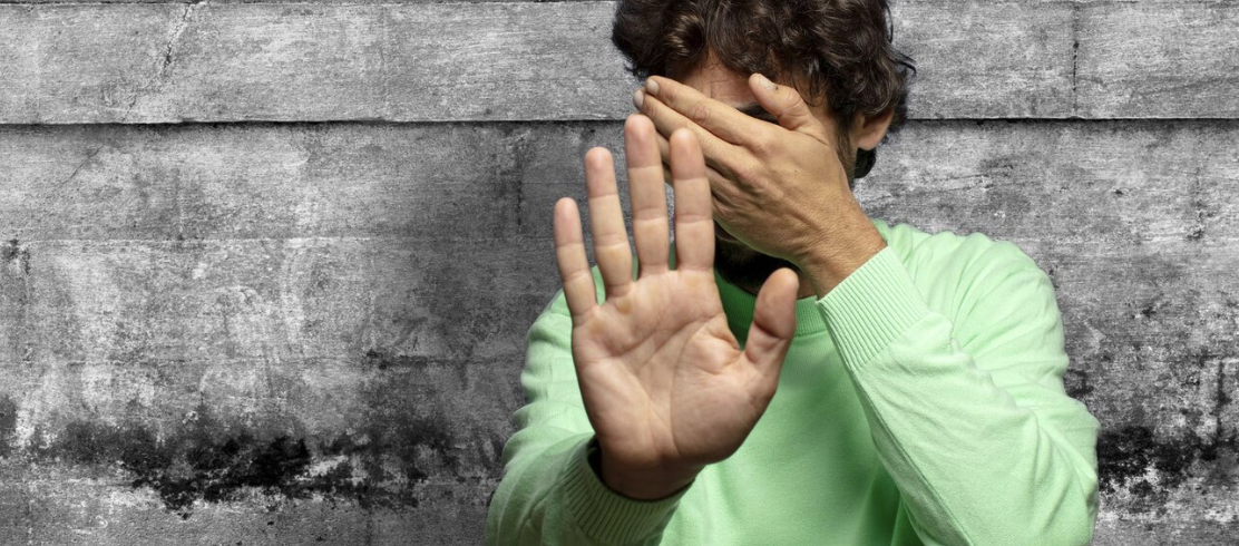 Nahaufnahme eines Jugendlichen vor grauer Wand, der sich schuetzend die Hand vors Gesicht haelt.