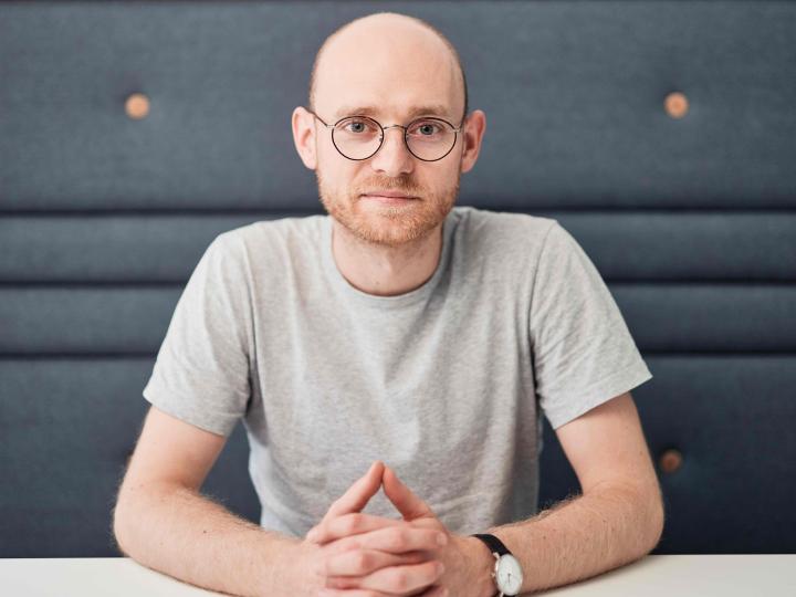 Portraitfoto David Alders, an einem Tisch sitzend, mit dunkelgrauem Hintergrund.