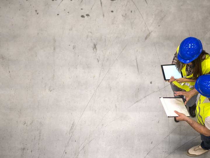 Zwei Arbeiter mit blauen Helmen und gelben Warnwesten auf einer Betonfläche aus der Vogelperspektive.