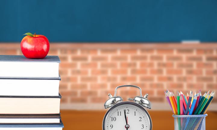 Stilleben Lehrerschreibtisch mit Buechern, Apfel, Wecker und Becher mit Buntstiften.