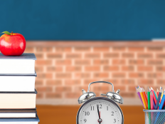 Stilleben Lehrerschreibtisch mit Buechern, Apfel, Wecker und Becher mit Buntstiften.