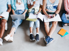 Gruppe junger Menschen auf dem Boden sitzend mit Buechern, Heften, Laptops und Smartphones.