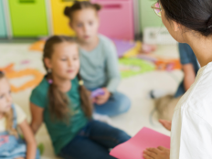 Drei Kindergartenkinder sitzen vor der Erzieherin und hören ihr zu.